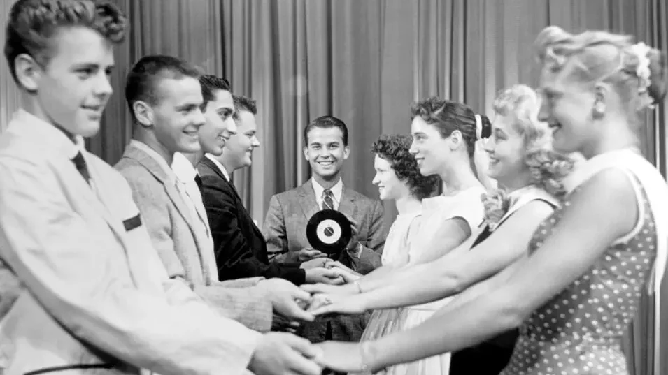 Teenagers dancing in a television studio in Philadelphia in the late 1950s, with studio lighting overhead and a live audience watching from bleachers.