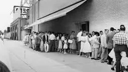 A black-and-white street scene in Southwest Philadelphia circa 1957, teenagers in plaid skirts and loafers walking toward a row house neighborhood on a sunny afternoon.