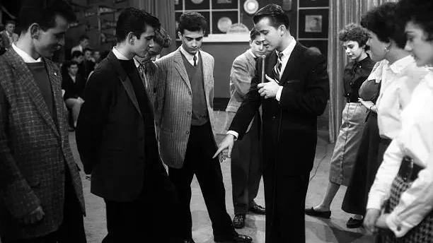 Teenage boys in jackets and ties and girls in dresses dance on a television studio floor under bright overhead lights. A camera operator stands at the edge of the frame.