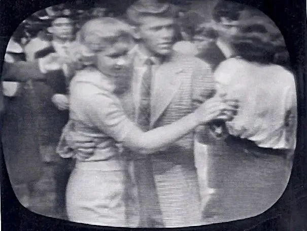 A teenage couple dances together on a television studio floor for the first time. The boy leads with quiet confidence, the girl follows. Studio lights illuminate them from above.