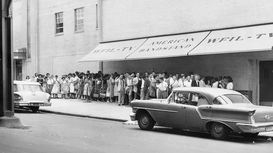 The exterior of a 1950s television studio building in Philadelphia, with a line of teenagers waiting outside on a city sidewalk.
