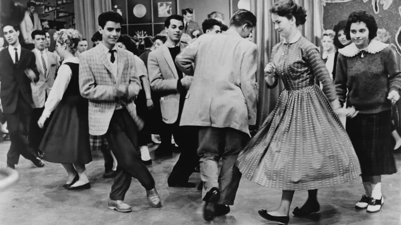 Interior of a 1950s television dance studio, teenagers in skirts and collared shirts dancing under studio lights, camera equipment visible at the edges of the frame.