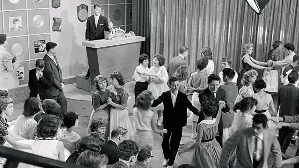 Pairs of teenage dancers move across a studio floor under bright television lighting. A young male performer stands at a microphone at the front of the room. The setting is a live television dance show studio, circa 1959. A visual recreation of the Bandstand studio floor in its peak years — where national youth culture was being assembled, broadcast, and quietly made permanent, one afternoon at a time.