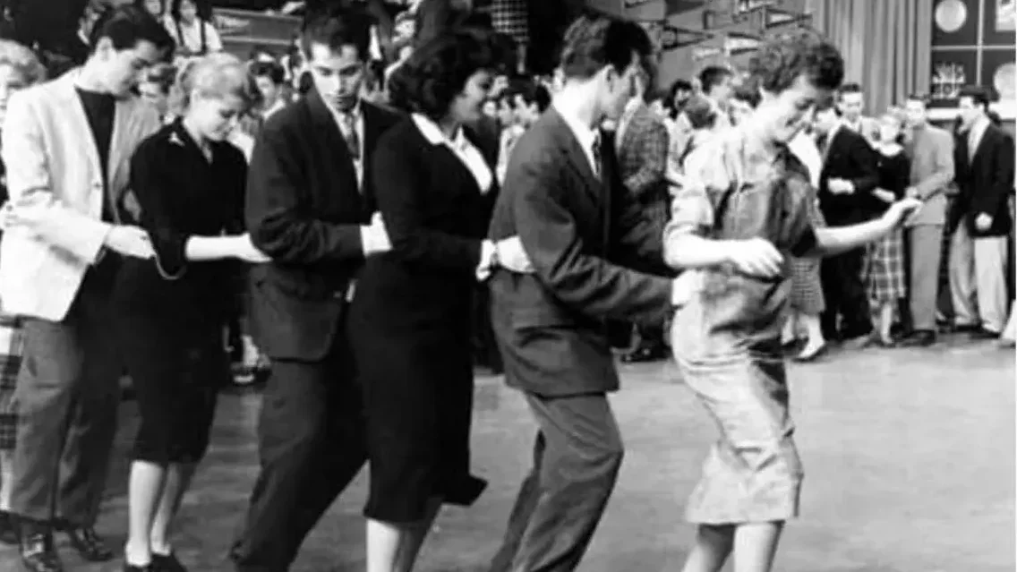 A wide shot of a television studio dance floor with teenage dancers in skirts and collared shirts. Camera equipment is visible at the edges. The floor is polished and reflects the overhead lights