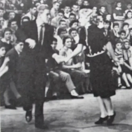 A young man and woman dance the Jitterbug on a television studio floor. The man leads with his right arm extended. Studio lights and other dancing couples are visible in the background.