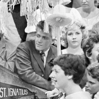 A young man and woman pose together at a public event. Both are neatly dressed. A crowd of teenagers is visible behind them, some reaching forward with papers for autographs.
