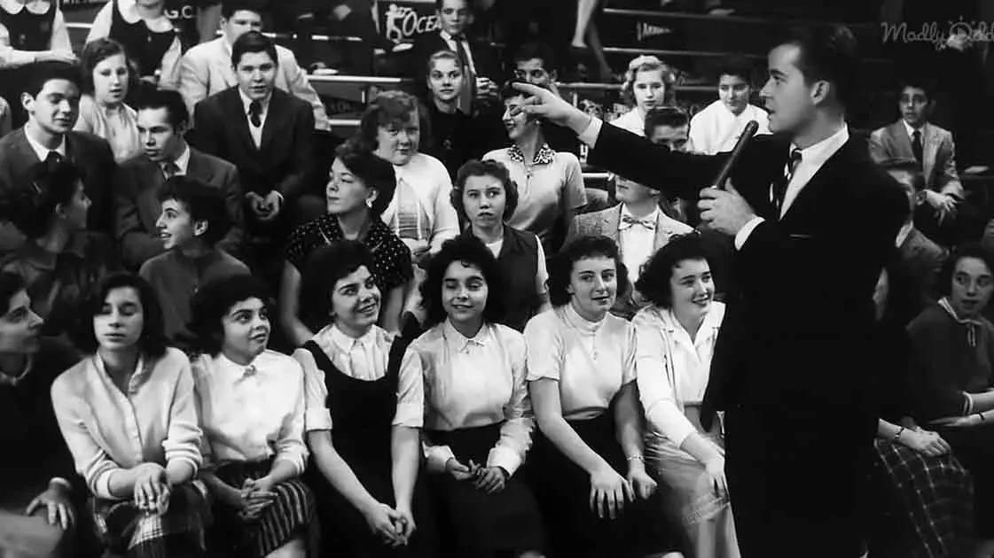 Teenagers dancing together on a 1950s television studio floor, wearing similar fashion styles and hairstyles, being filmed for a broadcast.