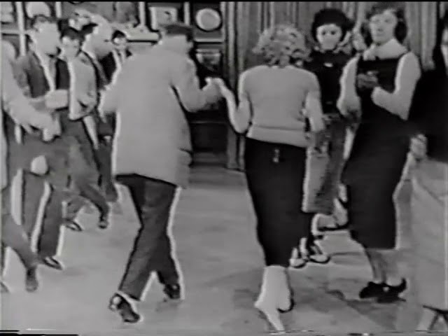 Two parallel lines of teenagers stand on a studio dance floor. Boys in suits on one side, girls in full skirts on the other. A couple moves through the center aisle between them.