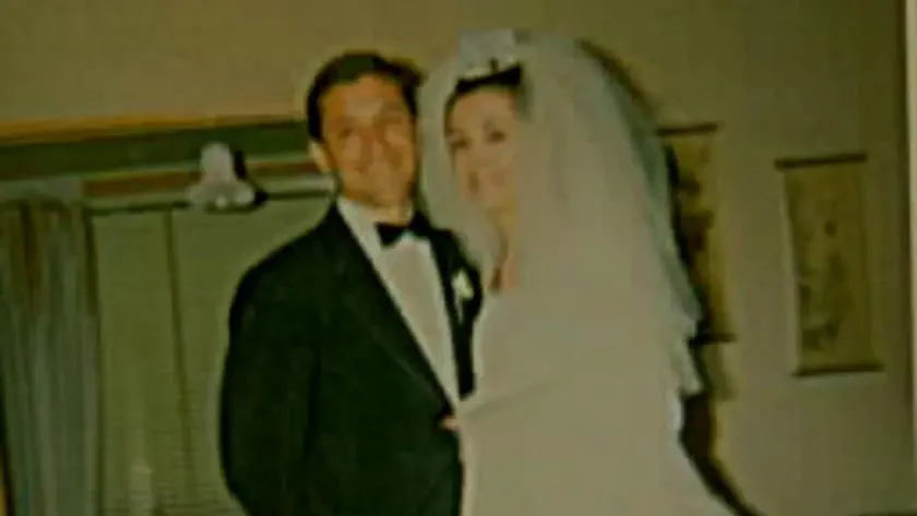 A color photograph of a bride and groom standing together indoors. The groom wears a black tuxedo with a bow tie and white boutonniere. The bride wears a full-length white wedding gown and veil with a small floral headpiece. Curtained windows and a framed picture are visible in the background.