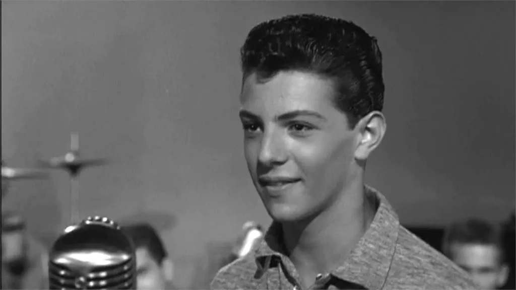 A young male performer stands at a studio microphone under bright stage lights. A studio audience of teenagers fills the background. The setting suggests a live television broadcast in the late 1950s. A visual recreation of a Saturday afternoon in 1957 — a studio floor in Philadelphia, a microphone, and a teenager about to become someone the whole country knew by name.
